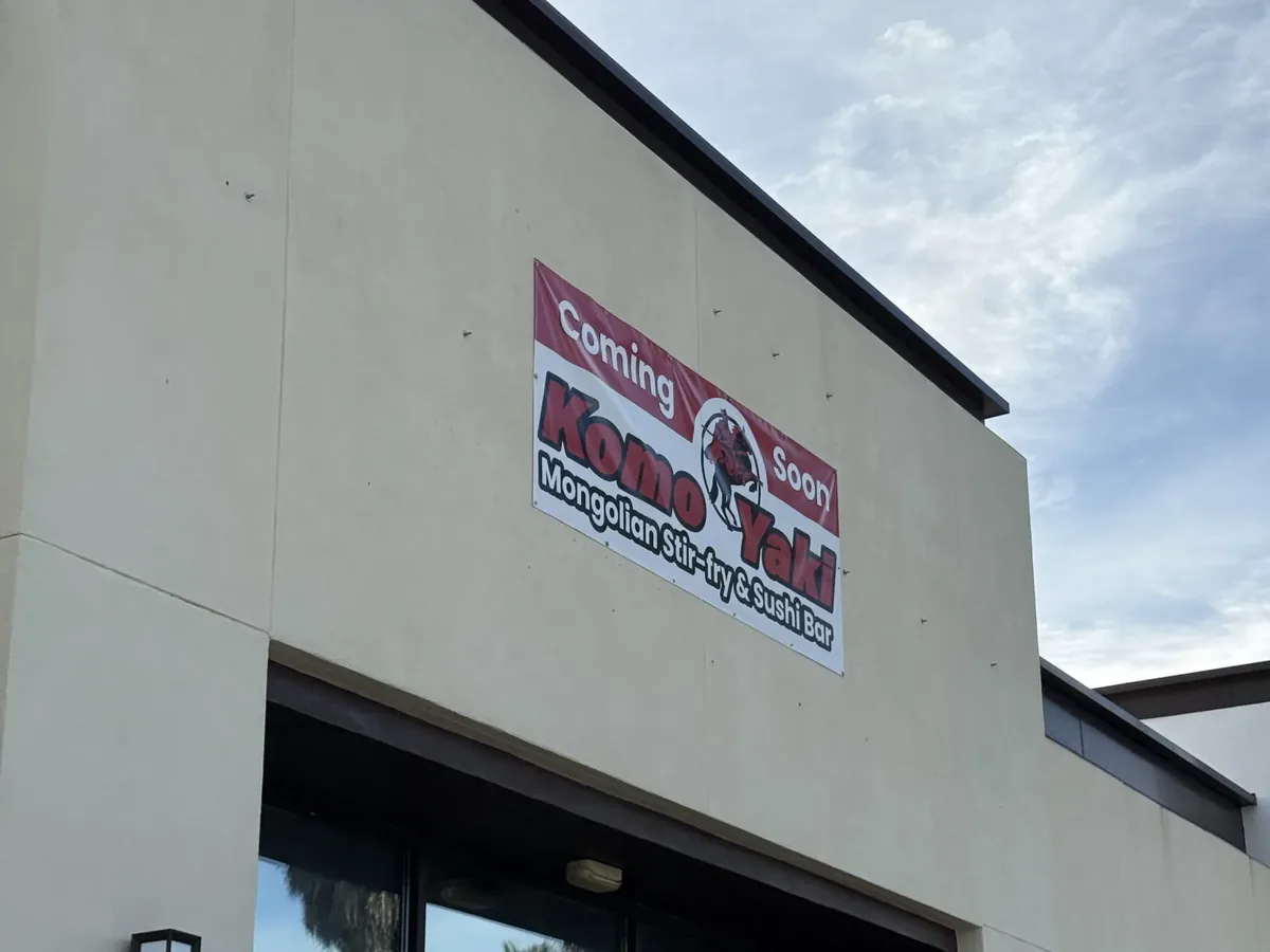 Red and white Coming Soon vinyl banner for Komo Yaki fastened to the restaurant's front wall above its glass entrance