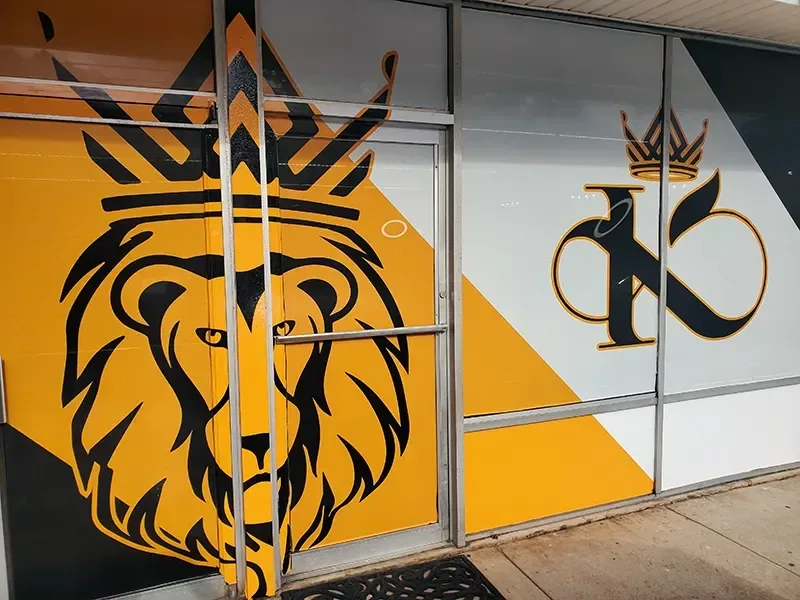 Vinyl lettering and window graphics covering a storefront's glass doors and windows: a crowned black lion's head and a crowned K logo on yellow, white and black panels